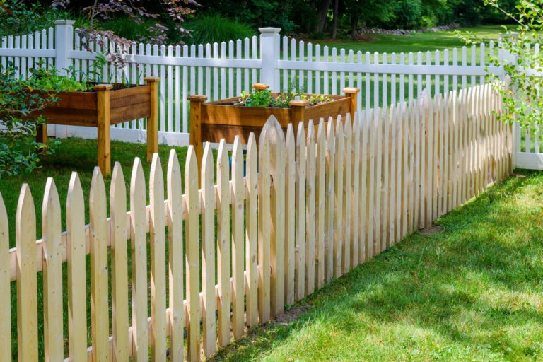 A wooden picket fence encloses a section of green lawn with raised wooden garden beds. A white picket fence borders the background, and trees and shrubs are visible beyond the fenced area.