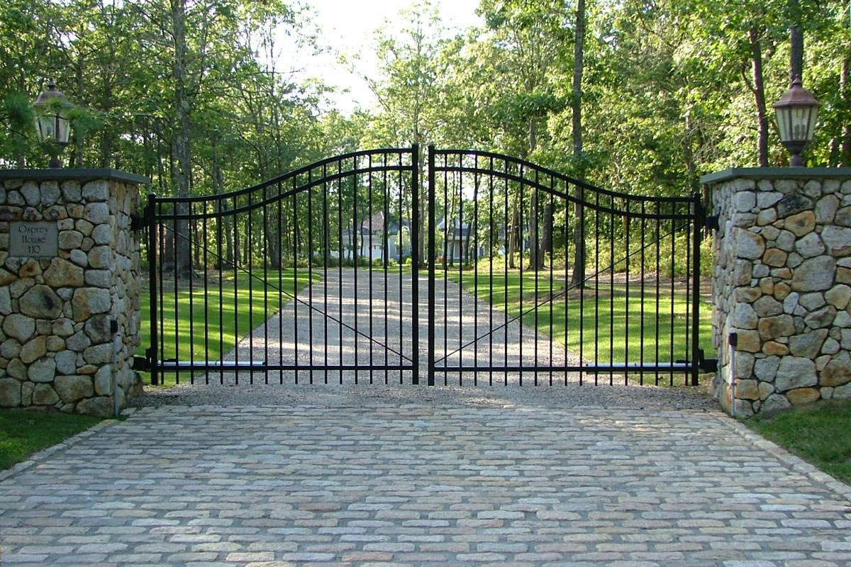 A black metal gate with decorative curves stands closed between two stone pillars with lanterns, blocking a cobblestone driveway that leads to a house surrounded by trees and grass.
