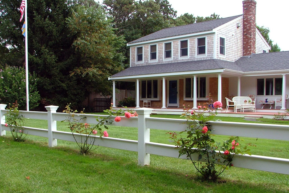 A two-story suburban house with white siding and a large front porch, a brick chimney, and a white fence with rose bushes in the yard. There is a flagpole on the lawn and trees in the background.