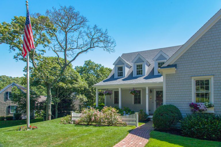 A two-story house with gray shingles, a front porch, and dormer windows sits on a manicured lawn with flower beds. An American flag is on a flagpole in the yard, and trees surround the property under a clear blue sky.