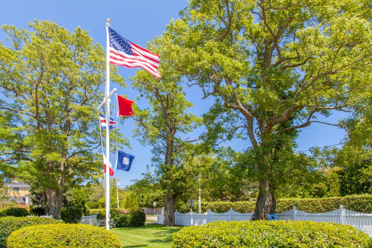 A flagpole with four flags, including the United States flag at the top, stands in a green, landscaped yard with large trees, a white fence, and shrubs under a clear blue sky.