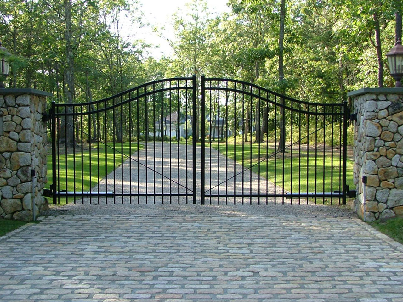 A black metal driveway gate is set between two stone pillars, with a cobblestone path leading to a tree-lined property in the background. The area is surrounded by green grass and trees.