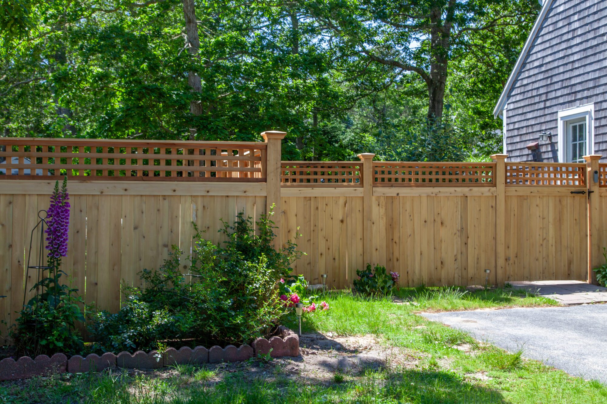 A wooden privacy fence with lattice panels runs along the edge of a yard. There are flowers and shrubs planted in front of the fence, and part of a house and driveway are visible on the right side. Trees are in the background.