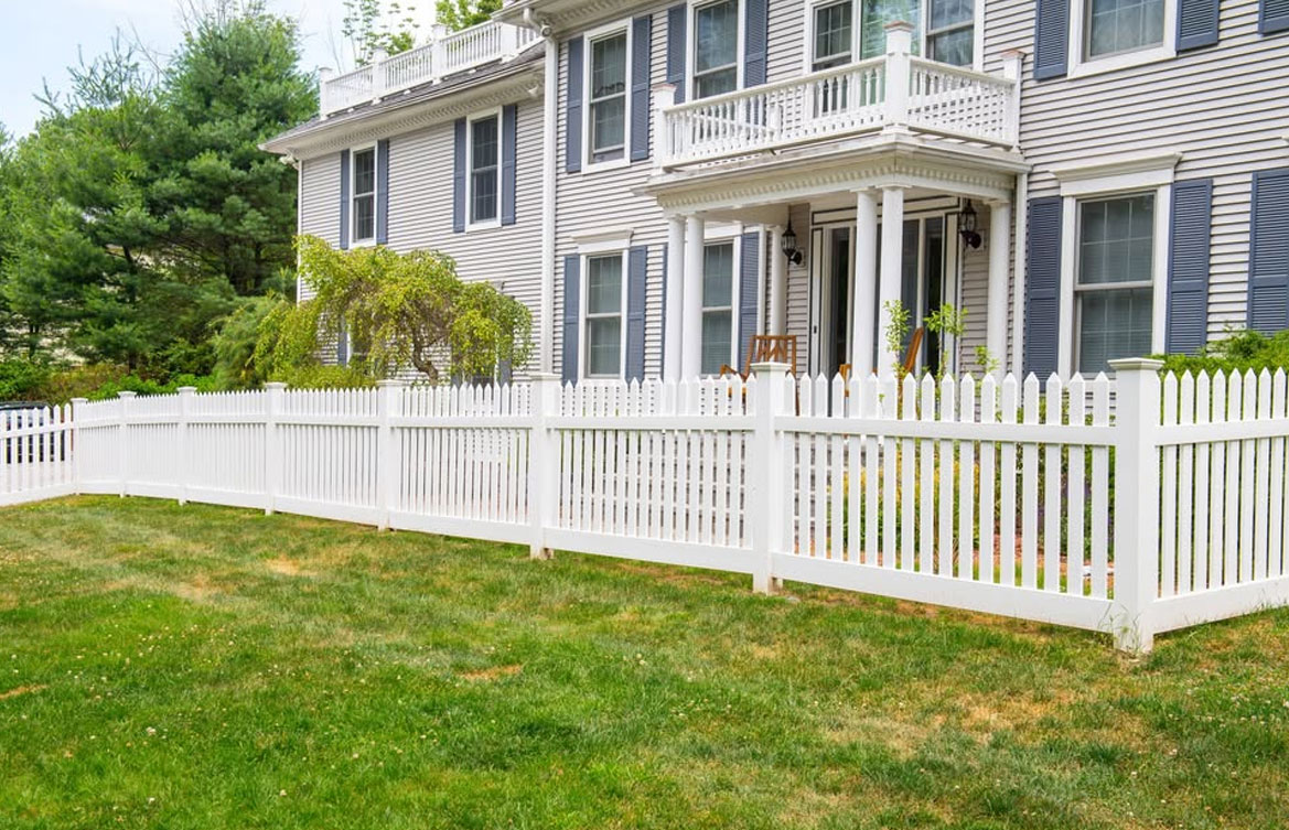 A white picket fence runs along the front yard of a large, two-story house with gray siding, blue shutters, and a small balcony above the main entrance. The lawn is green and well-kept, with shrubs near the fence.