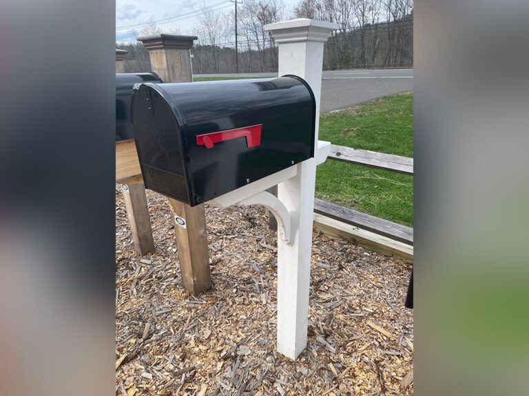 A black mailbox with a red flag mounted on a white wooden post, set near a road with a wooden fence and scattered wood chips on the ground. Trees and grass are visible in the background.