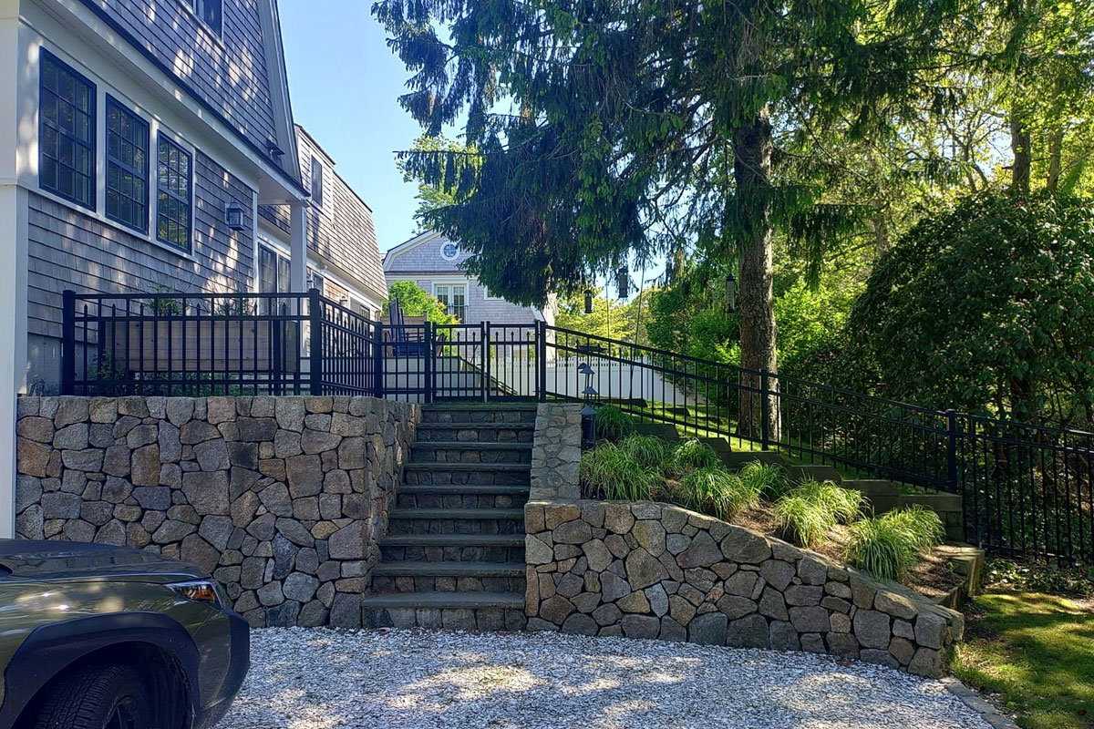 A stone staircase flanked by stone retaining walls leads up to a black metal fence and ramp beside a house. Green trees and shrubs surround the landscaped yard on a sunny day. A car is parked in a gravel driveway.