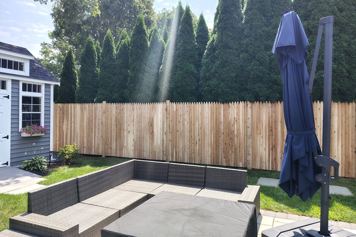 A backyard patio with wicker seating, a closed blue umbrella, wooden fence, green hedges, and sunlight streaming through the trees onto the grass.